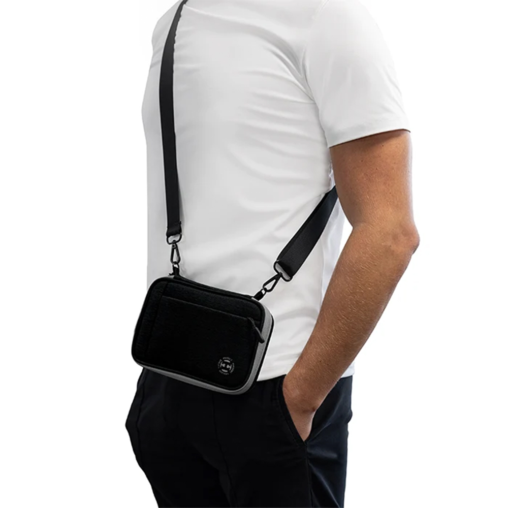 Une personne porte en bandoulière un étui à fléchettes noir « Harrows » Smart Case XL. Cet étui compact en tissu texturé, doté d’une bordure grise, est muni d’une sangle réglable. T-shirt blanc, une main dans la poche du pantalon.