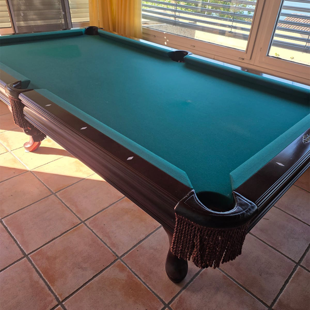An elegant snooker table with emerald-green baize and a dark wooden frame, featuring striking corners with brown leather fringing. It stands on terracotta-coloured tiles, bathed in sunlight streaming through the window with blinds.