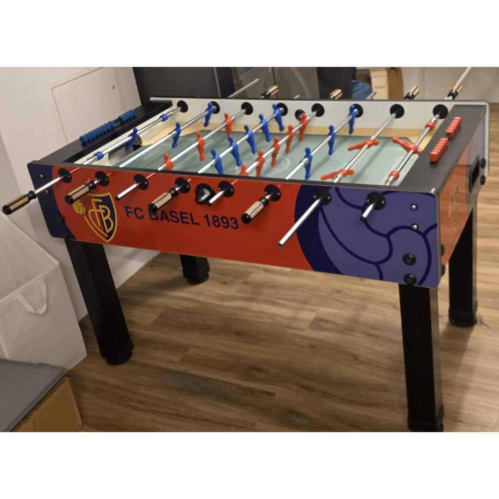 Une table de baby-foot rouge, bleue et orange, dotée de pieds noirs, est posée sur un parquet. Des figurines bleues et rouges sont fixées sur les barres. Sur les côtés, on peut lire « FC BASEL 1893 ». Bien éclairée, dans un intérieur.