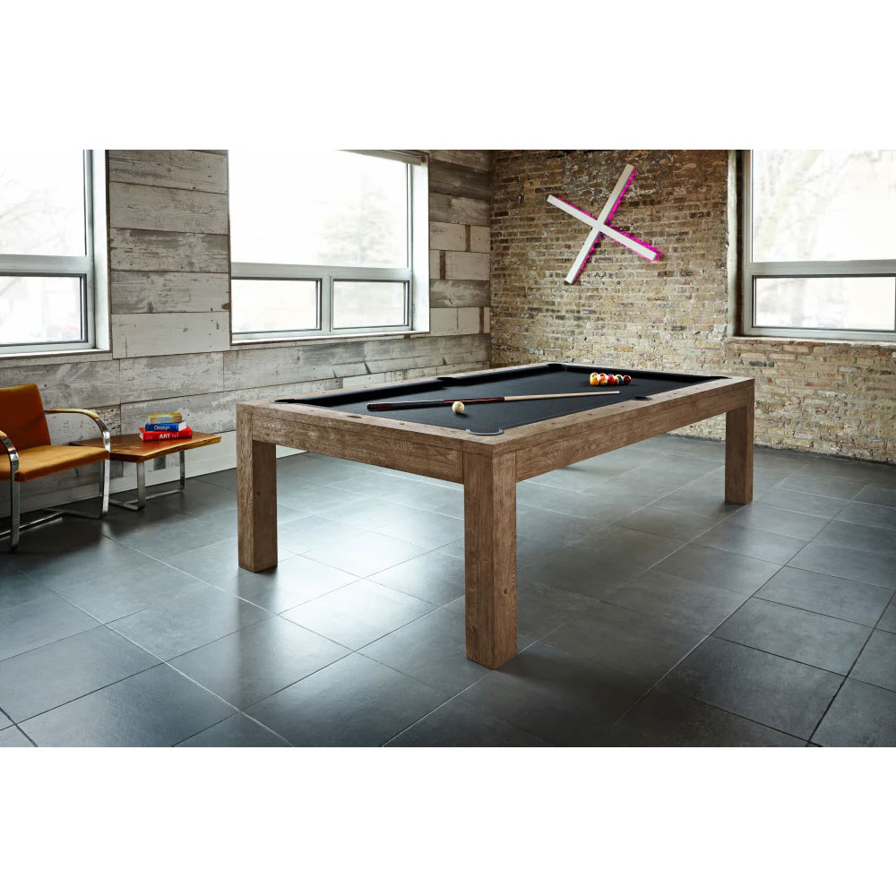 A modern pool table— Brunswick Sanibel—in a Rustic Dark Brown wood finish with black cloth. Pool balls and a cue rest on the table. The table stands in a bright room with large windows, brick walls and wood panelling.
