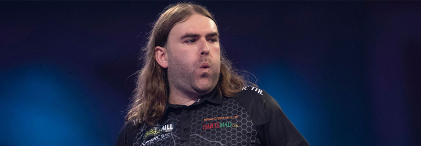 Un homme aux longs cheveux bruns et à la barbe de trois jours regarde à droite, concentré, la bouche légèrement entrouverte. Il porte un polo foncé à motifs avec des logos de sponsors. Arrière-plan flou bleu sombre, évoquant sport ou scène.