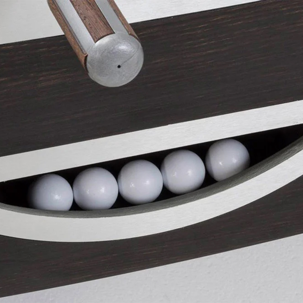Gros plan sur une table de baby-foot moderne ou jeu similaire. Une tige avec manche en bois et embout argenté dépasse en haut. Dessous, plateau en bois foncé à bandes blanches. Cinq balles blanches brillent dans une rainure.