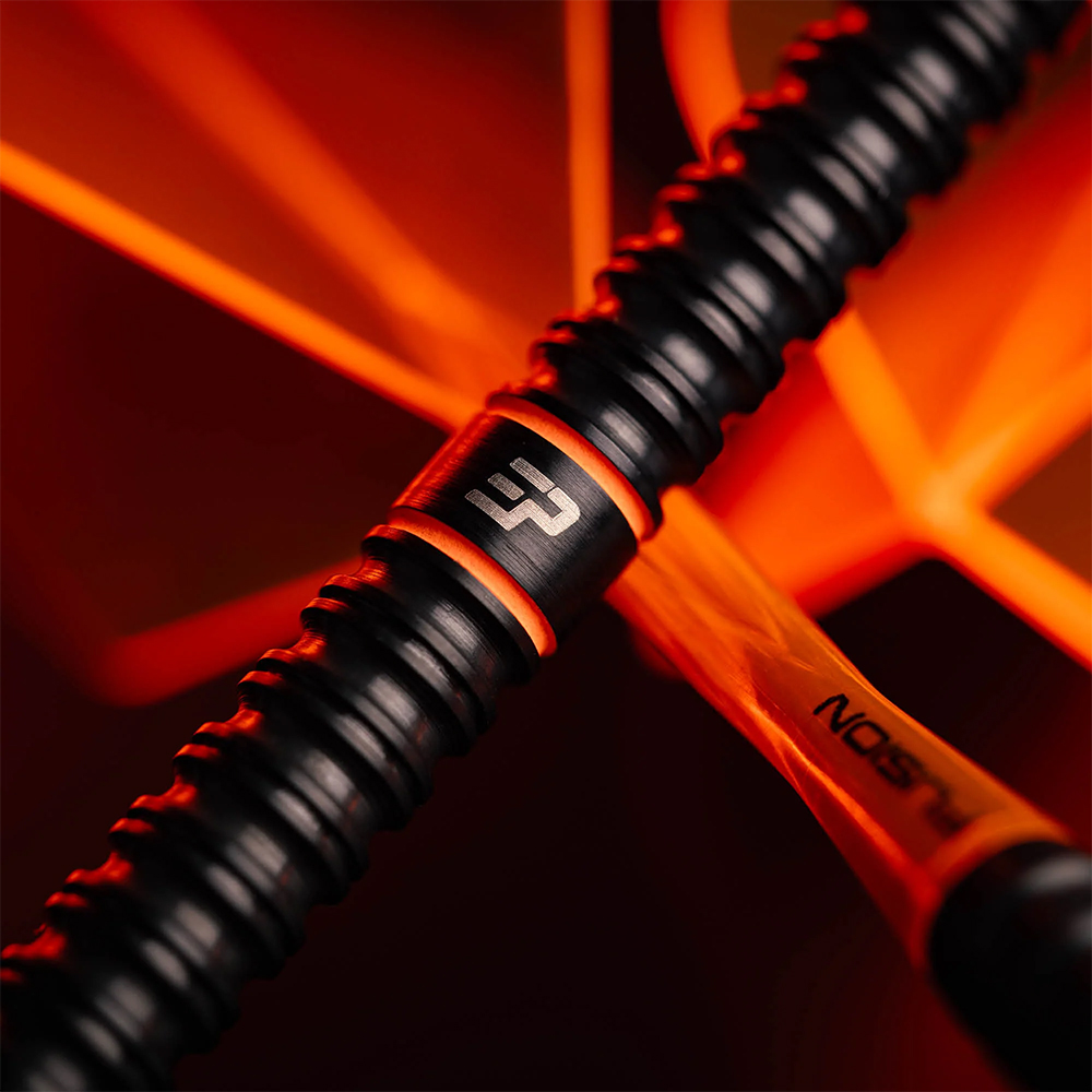 Nahaufnahme eines schwarzen Steeldart-Barrels mit feiner Rillenstruktur für Griffigkeit. Ein glattes Mittelstück trägt das silberne EP-Logo, umrahmt von orangefarbenem Lichtschimmer. Im Hintergrund ein orange-gelber Fusion-Flight.