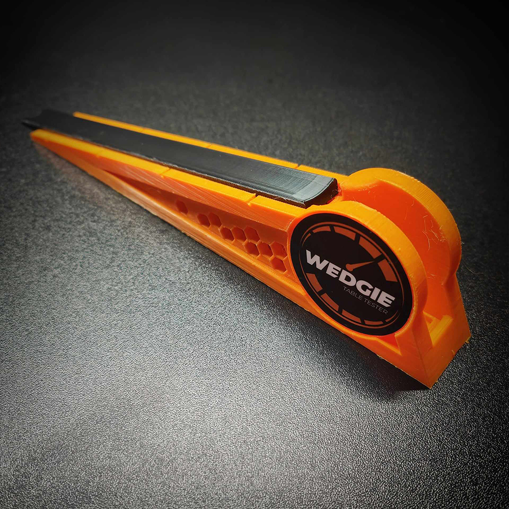 Un testeur de table de billard Wedgie orange, en forme de coin, repose sur un fond sombre. Dessus noir lisse avec une étiquette ronde « WEDGIE TABLE TESTER ». Côtés à motif alvéolé, de construction légère, destiné au contrôle des tables.