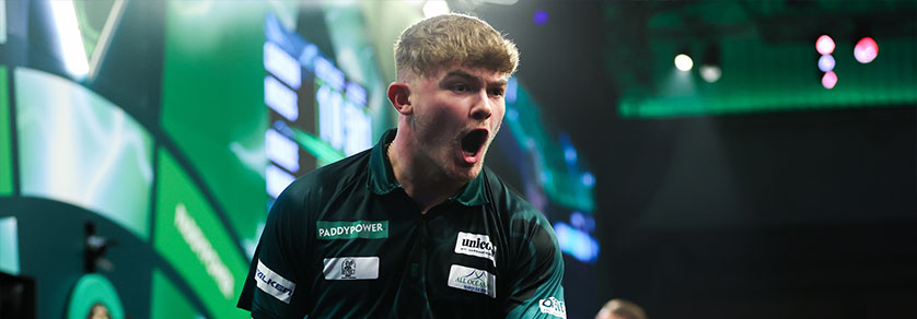 Un jeune homme aux cheveux blonds exulte, la bouche grande ouverte, et regarde sur le côté. Il porte un polo vert foncé orné de logos de sponsors, comme Paddy Power. L'arrière-plan flou, parsemé de lumières vertes, suggère une salle de sport.