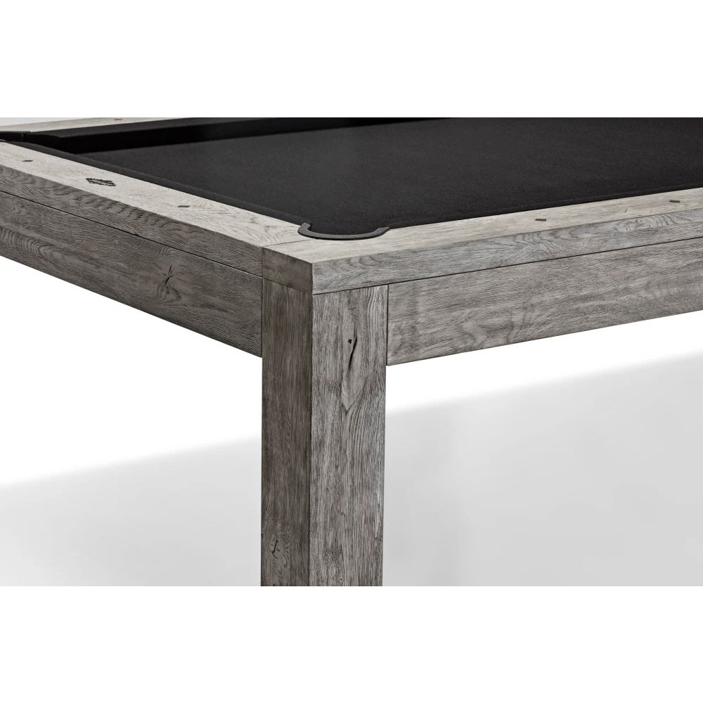 Close-up view of the corner of a modern pool table. Frame and legs made of wood with a rustic grey finish, featuring visible grain and slight indentations. Playing surface covered with black cloth. A dark, flat corner pocket is visible.