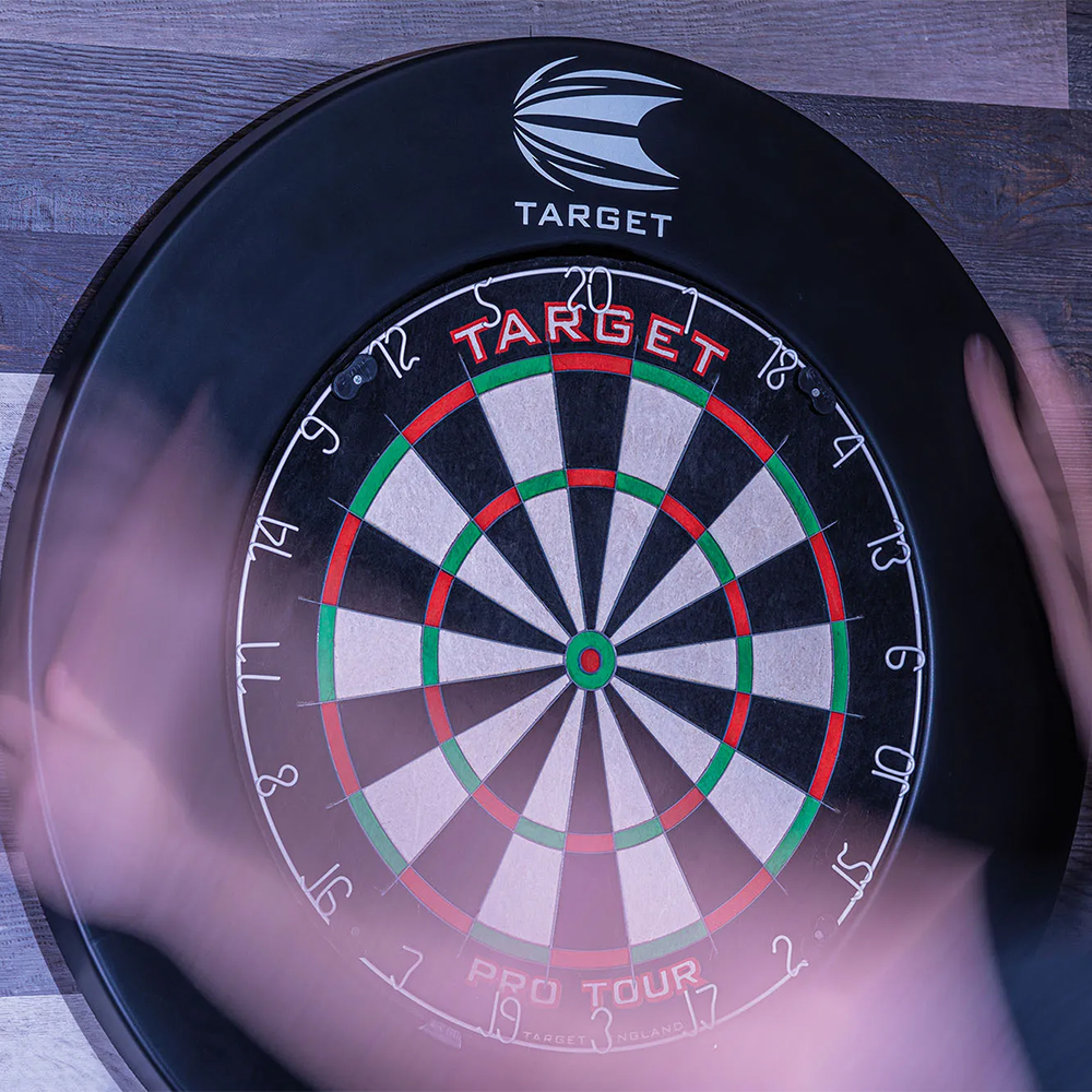 A black ‘ Target ’ dartboard with colourful segments in black, white, red and green hangs on a wooden wall. The numbers 1 to 20 are visible around the edge. In the foreground, there is a blurred movement, presumably of a person playing.