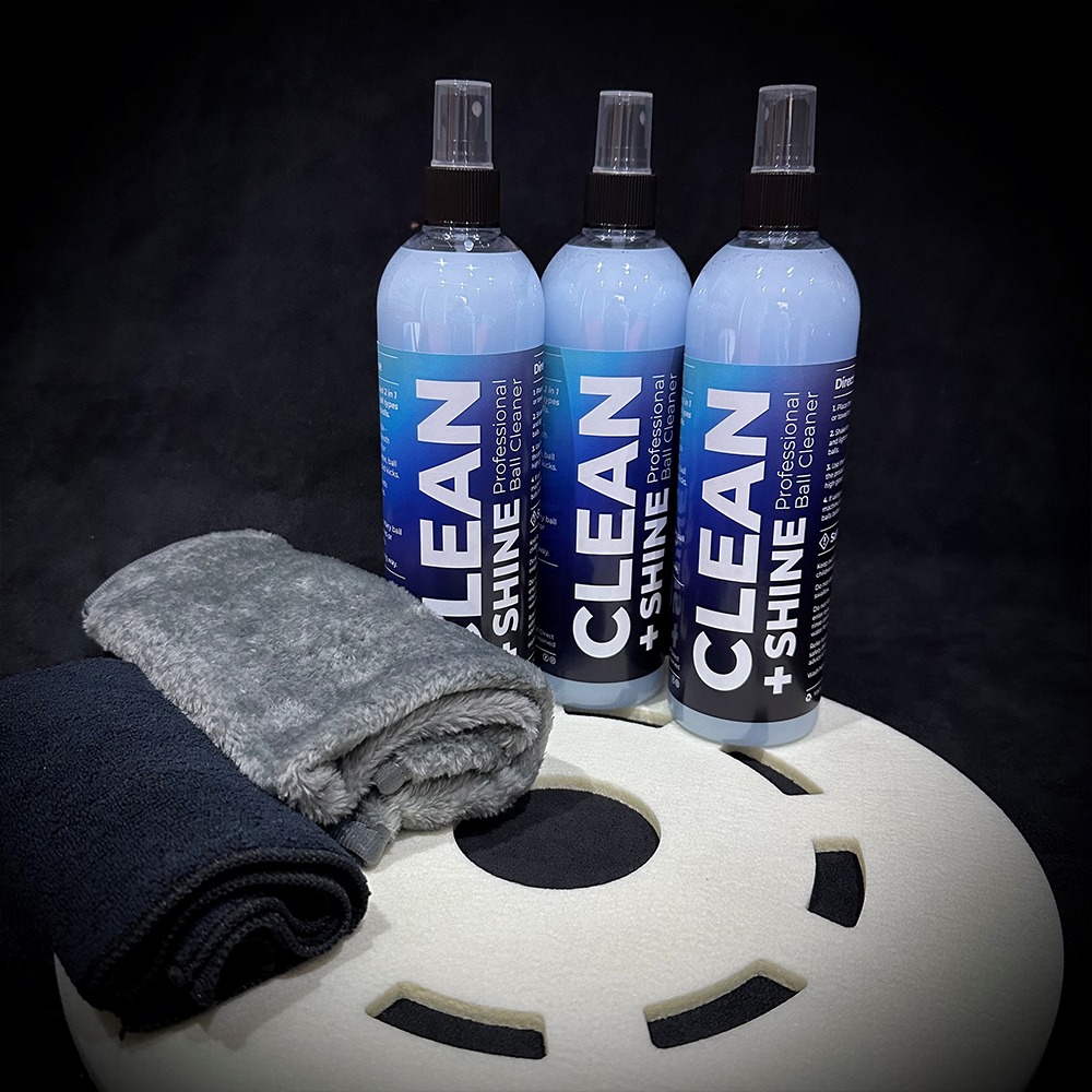 Drei hellblaue Sprühflaschen „CLEAN + SHINE Professional Ball Cleaner“ vor schwarzem Hintergrund. Davor graues, flauschiges Handtuch und dunkles Mikrofasertuch. Vorn rundes, weisses Pad mit schwarzen Aussparungen.