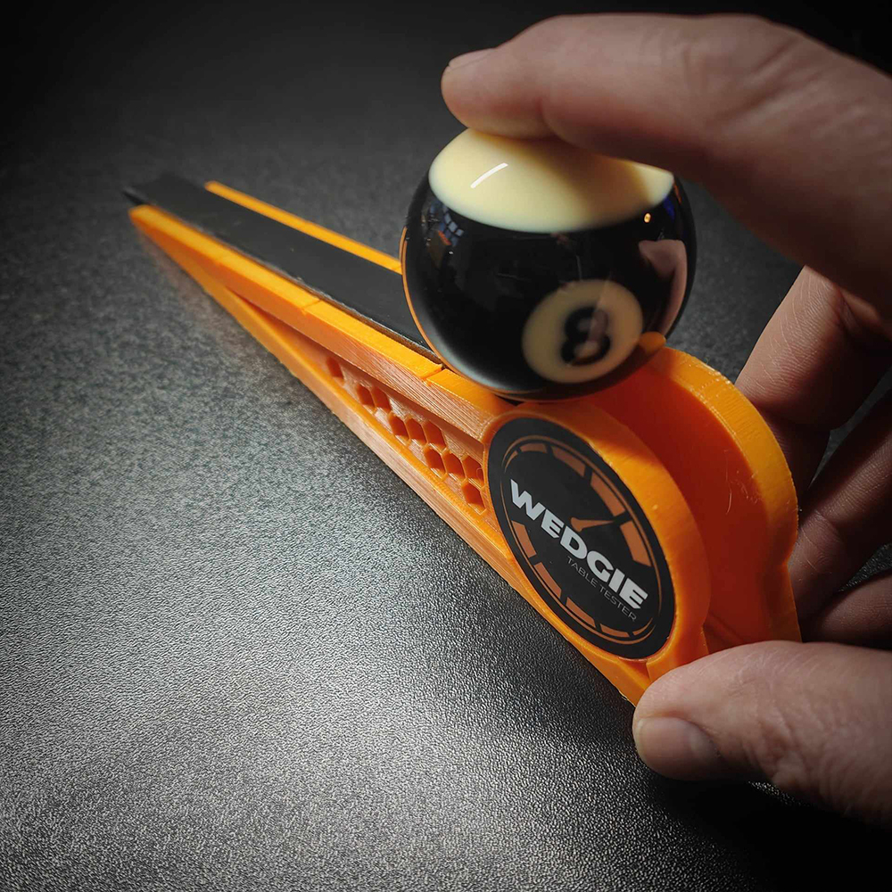 A hand is holding an orange Wedgie billiard table tester. A black 8-ball lies on the slightly sloping surface. The device tests the speed and accuracy of billiard tables; the ‘WEDGIE TABLE TESTER’ logo is visible.