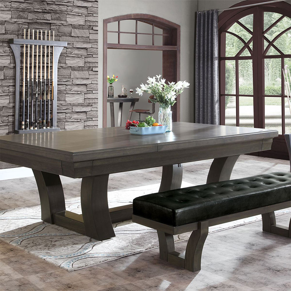 Salle à manger avec grande table en bois gris et banquette en cuir noir. Vase de fleurs blanches et fruits sur la table. Mur en pierre avec queues de billard, fenêtre en arc, bar au fond.