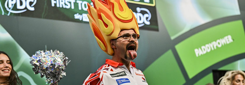 Un homme à lunettes, moustache et chapeau jaune-orange à flammes tire la langue. Il porte une chemise blanche avec logos de sponsors et regarde légèrement à droite. En arrière-plan : murs verts « PDC/PADDYPOWER », femme au pompon.