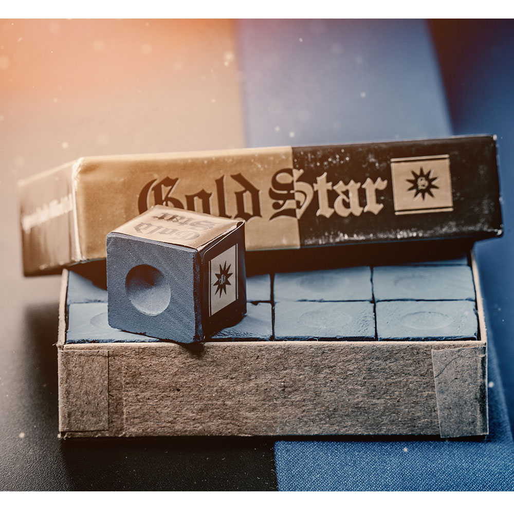 Offene, leicht verwitterte Pappschachtel mit mehreren quadratischen, himmelblauen Billardkreiden. Eine Kreide liegt auf dem Karton, zeigt eine runde Vertiefung und das Gold Star Logo. Warmes Licht.