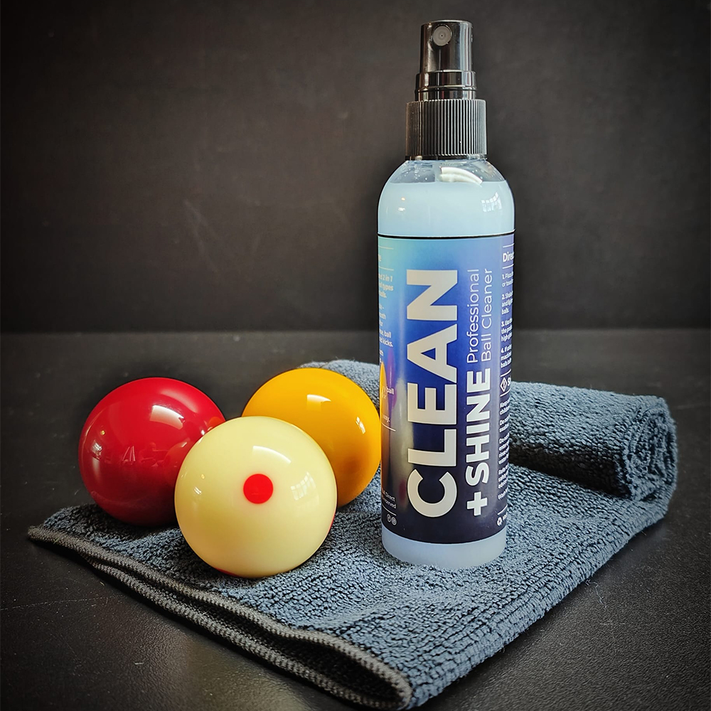 Flacon « Clean + Shine Professional Ball Cleaner » sur chiffon microfibre gris foncé. À côté, trois boules de billard brillantes : rouge, jaune et blanche à point rouge. Promet des surfaces lisses et ultra-brillantes.