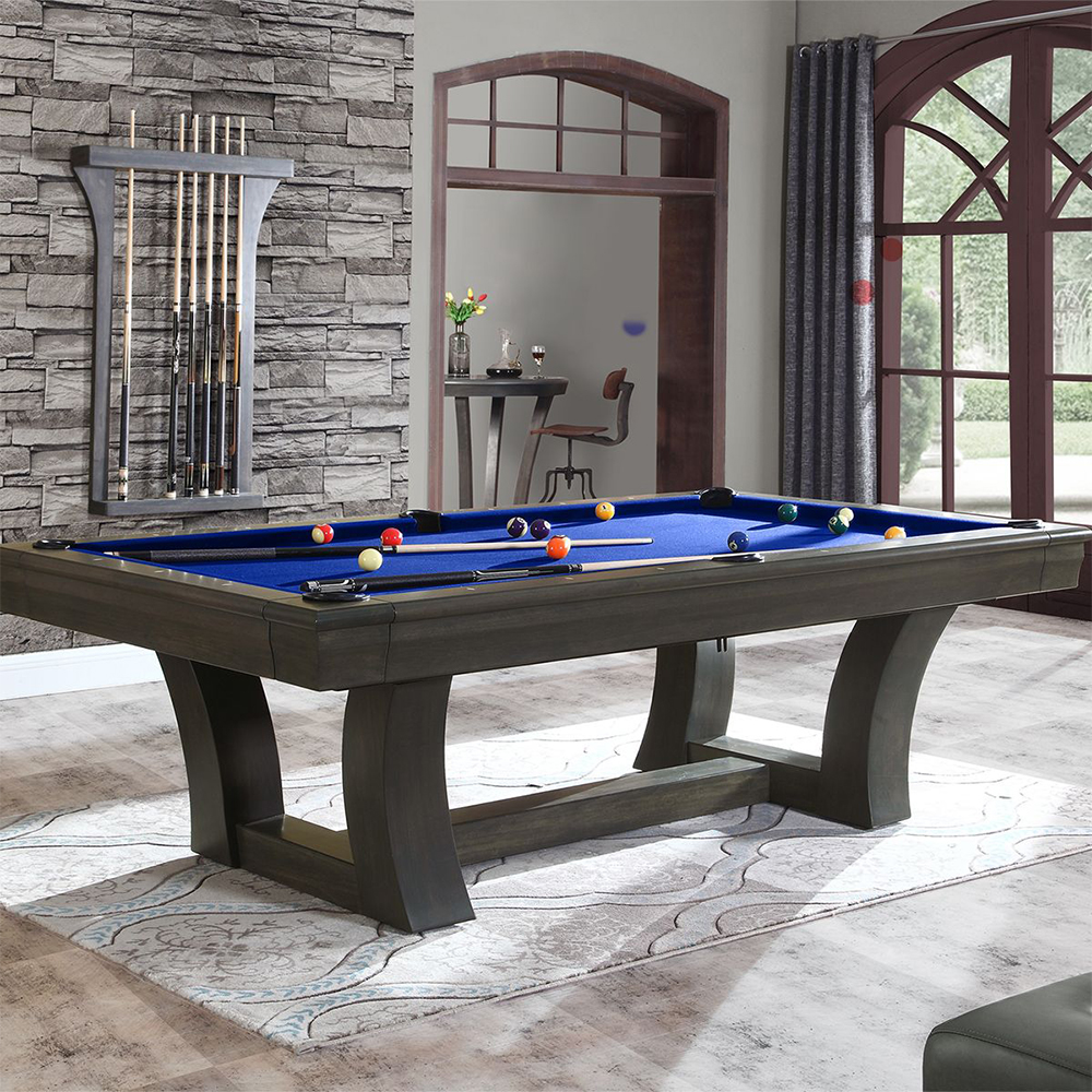 Salle de billard moderne avec une table élégante gris foncé et un feutre bleu. Des boules colorées et des queues sont à disposition. Un porte-queues est fixé au mur en pierre. À l'arrière-plan : une pièce avec fenêtre en arc et table de bar.