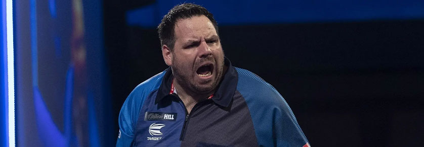 Un homme barbu aux cheveux foncés, vêtu d’un polo bleu-gris orné de logos, ouvre grand la bouche et crie ou jubile. Expression intense, regard tourné vers la droite. Arrière-plan sombre et flou avec des touches de lumière bleue, événement sportif.