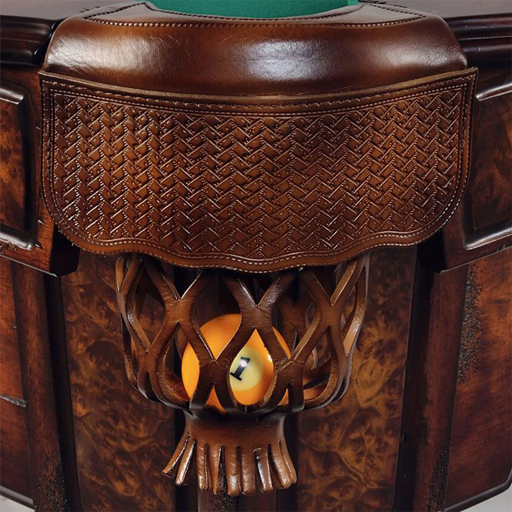 Zoom avant sur un sac de billard en cuir marron foncé. Le rabat est finement gaufré d'un motif tressé et contient une boule de billard n° 1 rayée jaune et orange. La table est en bois noble veiné.