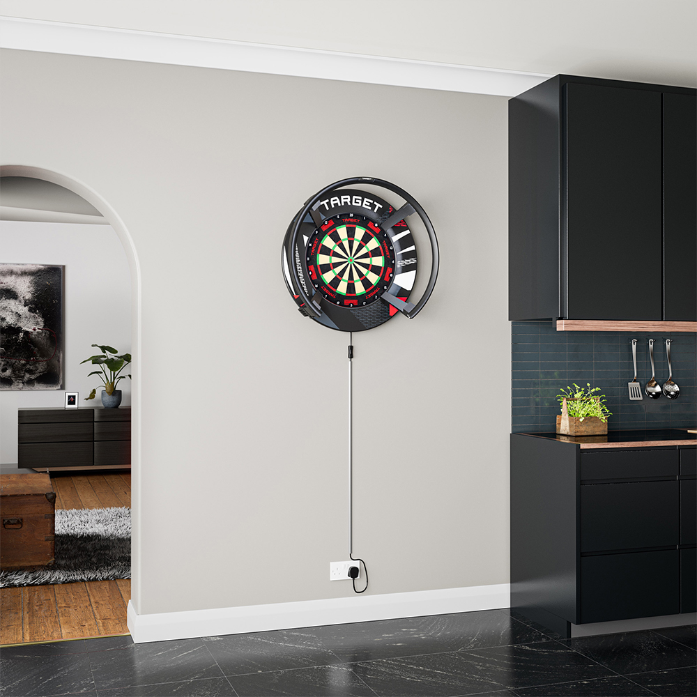 Un anneau de protection noir « Target » entoure une cible de fléchettes éclairée, fixée à un mur gris clair dans un salon moderne. L'anneau protège le mur et intègre un éclairage ; le câble est branché. À droite : cuisine noire.