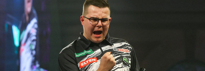 Un jeune joueur de fléchettes portant des lunettes et un polo noir et blanc exulte, la bouche grande ouverte et le poing serré. On aperçoit sur son polo les logos « PADDYPOWER », « ELTEN » et « REDDRAGON », sur un fond sombre et flou.