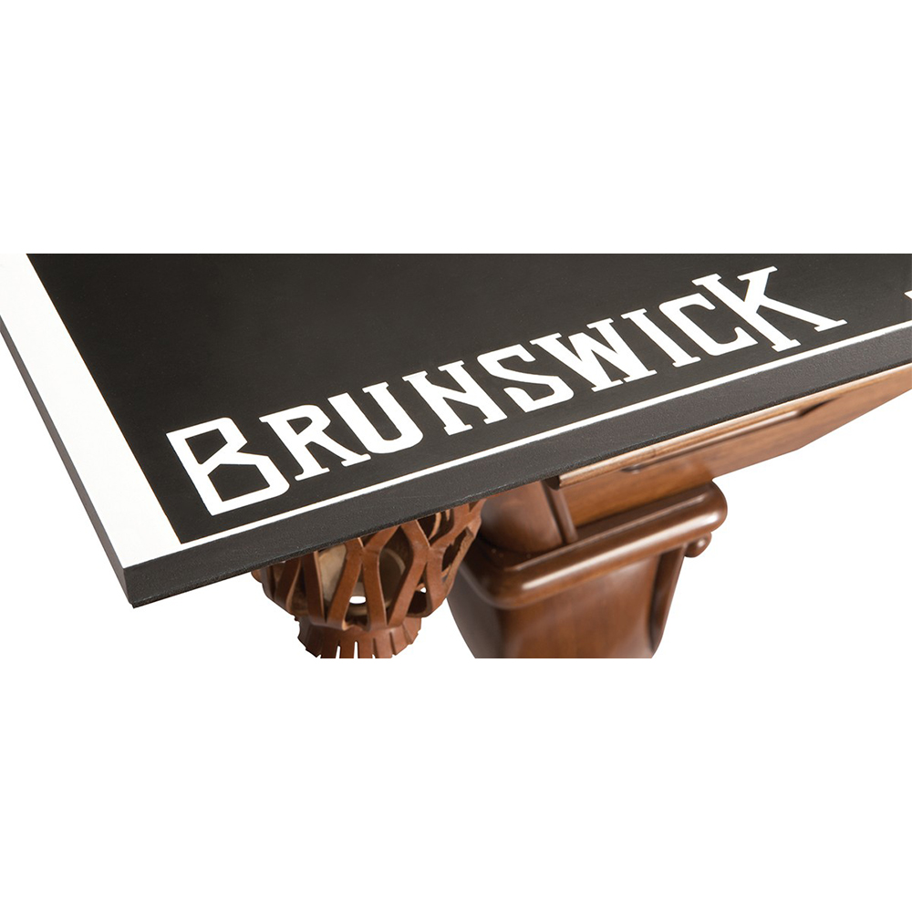 Zoom avant sur le coin d’une table de billard à surface de jeu noir mat, fine ligne blanche sur le pourtour. Le nom de la marque « Brunswick » apparaît en grosses lettres blanches. En dessous, panier de boules en bois clair.