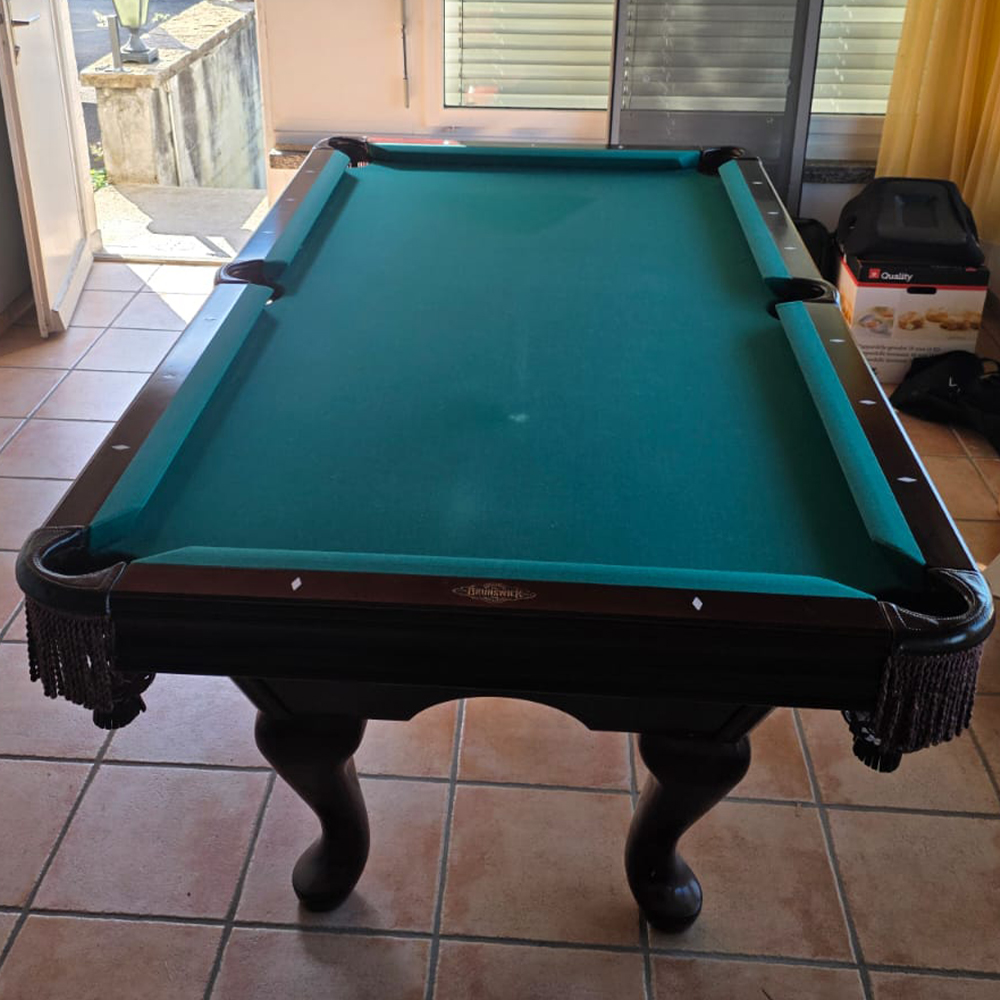 A large billiards table with dark green baize and a brown wooden frame, tassels on the pockets, standing on a light brown tiled floor. In the background: an open passageway leading outside, a stone staircase, a lantern and a window with blinds.