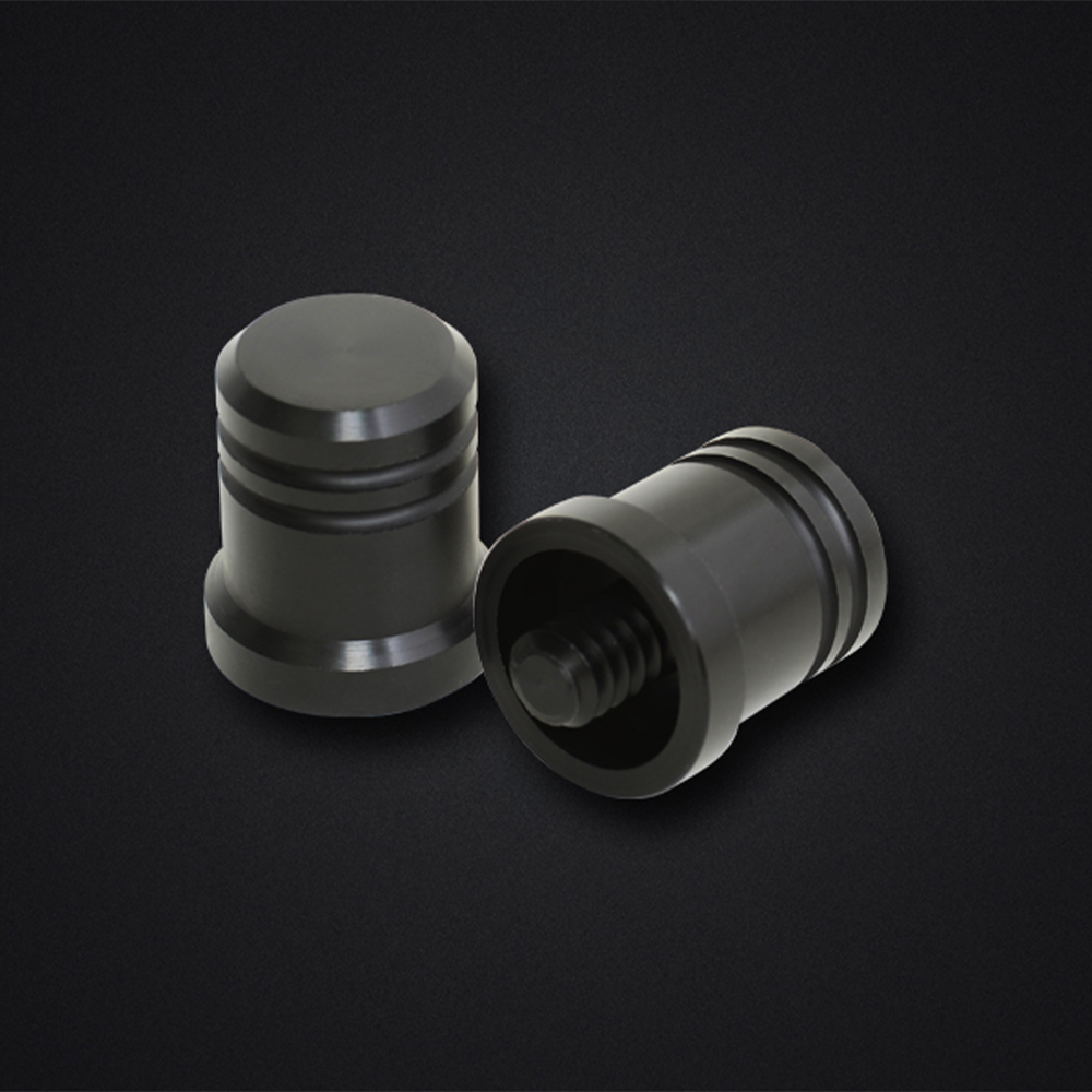 Deux capuchons métalliques cylindriques identiques, noir mat, sur fond sombre légèrement texturé. L'un est vertical, face supérieure lisse et deux rainures sur le pourtour. L'autre est incliné, avec ouverture et filetage intérieur.