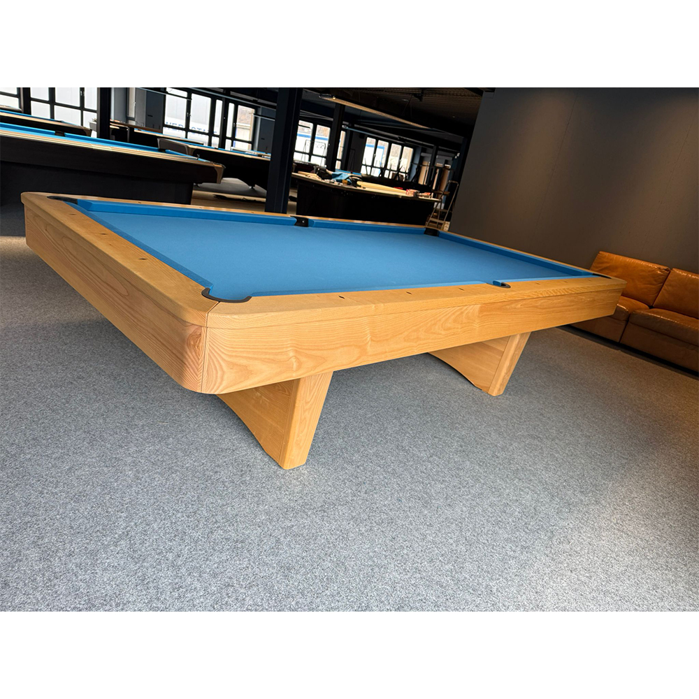Table de billard Kairos avec un cadre en bois clair et un tapis bleu vif. Ses pieds robustes et incurvés en bois massif lui confèrent un aspect élégant. À l'arrière-plan, d'autres tables et un canapé en cuir marron.