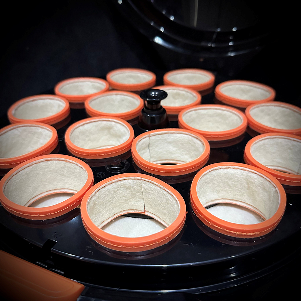 Nahaufnahme des Innenraums einer Billard-Kugelpoliermaschine mit 16 runden Reinigungsbechern. Jeder Becher hat einen orangefarbenen Rand und ist mit weicher, weißer Wolle ausgekleidet. Kreisförmig zum Polieren.