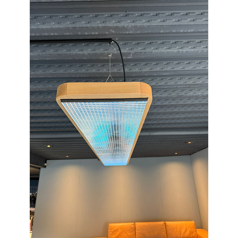 Suspension allongée avec structure en bois clair et abat-jour strié transparent, diffusant une lumière froide bleutée. Suspendue à un plafond métallique gris, elle éclaire la pièce avec la table de billard américain Kairos.