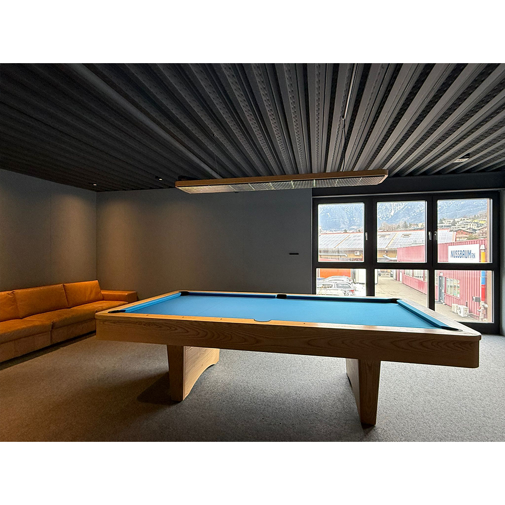 Table de billard « Kairos » en bois massif clair, tapis bleu, dans une pièce moderne. À gauche, long canapé orange sous une lampe en bois. Grandes fenêtres avec vue montagnes et ville. Plafond sombre, moquette grise.