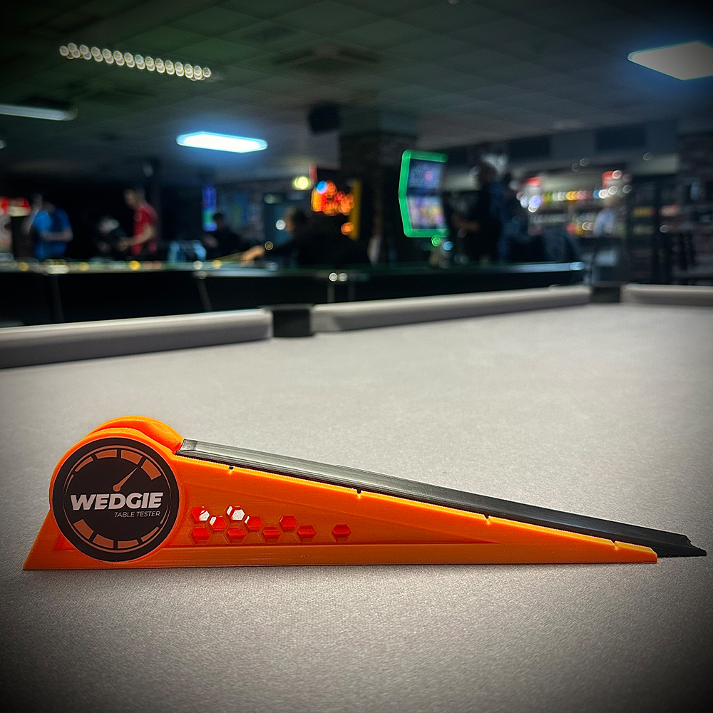 An orange and black Wedgie Billiard Table Tester lies on grey billiard cloth. The wedge-shaped device, bearing the ‘Wedgie Table Tester’ logo, is ready to test table speed and precision. In the background are more tables and people.