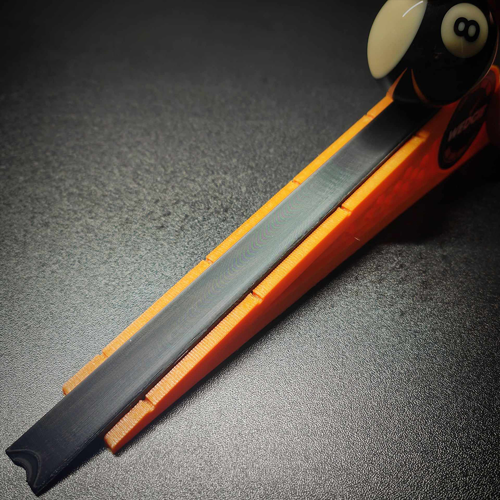 An orange and black Wedgie pool table measuring device lies on a dark surface. A black 8-ball rests at the top of the orange frame, ready to roll down the black measuring rail. For testing speed and accuracy.