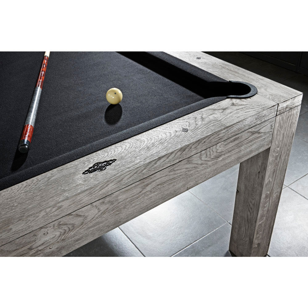 Brunswick Sanibel pool table in Rustic Grey with visible wood grain in a weathered finish. A white ball and a cue rest on black baize. Flat corner pockets emphasise the modern design.