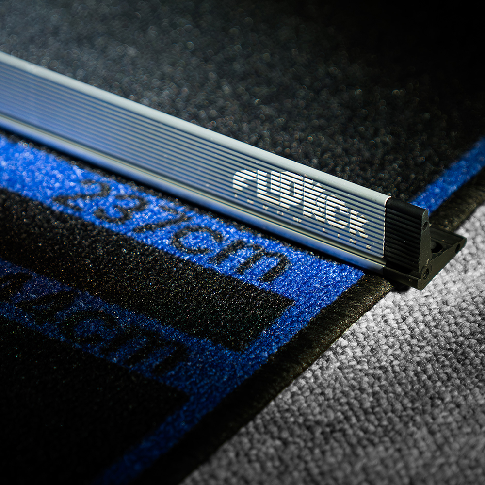 Gros plan sur l'« Oche » FlipKick argenté de Target, une ligne de lancer surélevée pour joueurs de fléchettes. Le profilé en aluminium usiné, marqué « FLIPKICK », repose sur un tapis noir sur moquette grise.