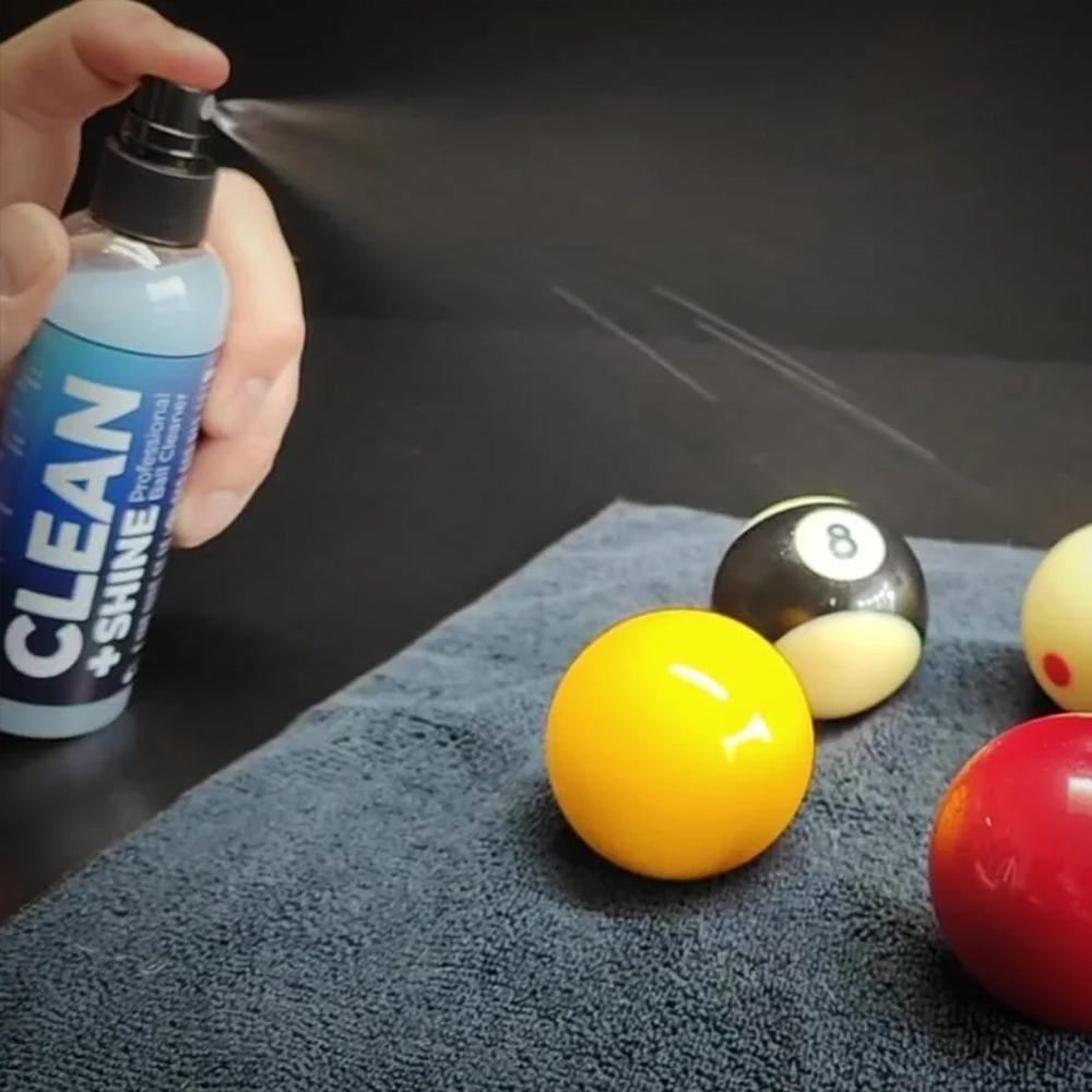 Une main vaporise du « CLEAN + SHINE Professional Ball Cleaner » avec un flacon transparent sur un chiffon en microfibre gris foncé. Des boules de billard sont posées dessus : une jaune, une noire (8) et une rouge.