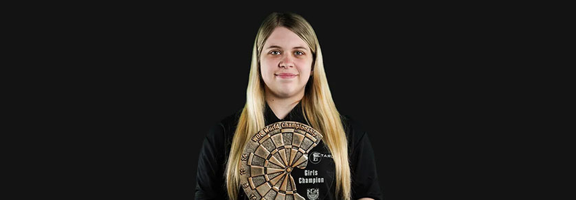 Eine junge Frau mit langen blonden Haaren und leichtem Lächeln steht vor dunklem Hintergrund. Sie hält eine runde Trophäe wie eine Dartscheibe mit "WDC World Championship" und "Girls Champion". Sie trägt ein schwarzes Hemd.
