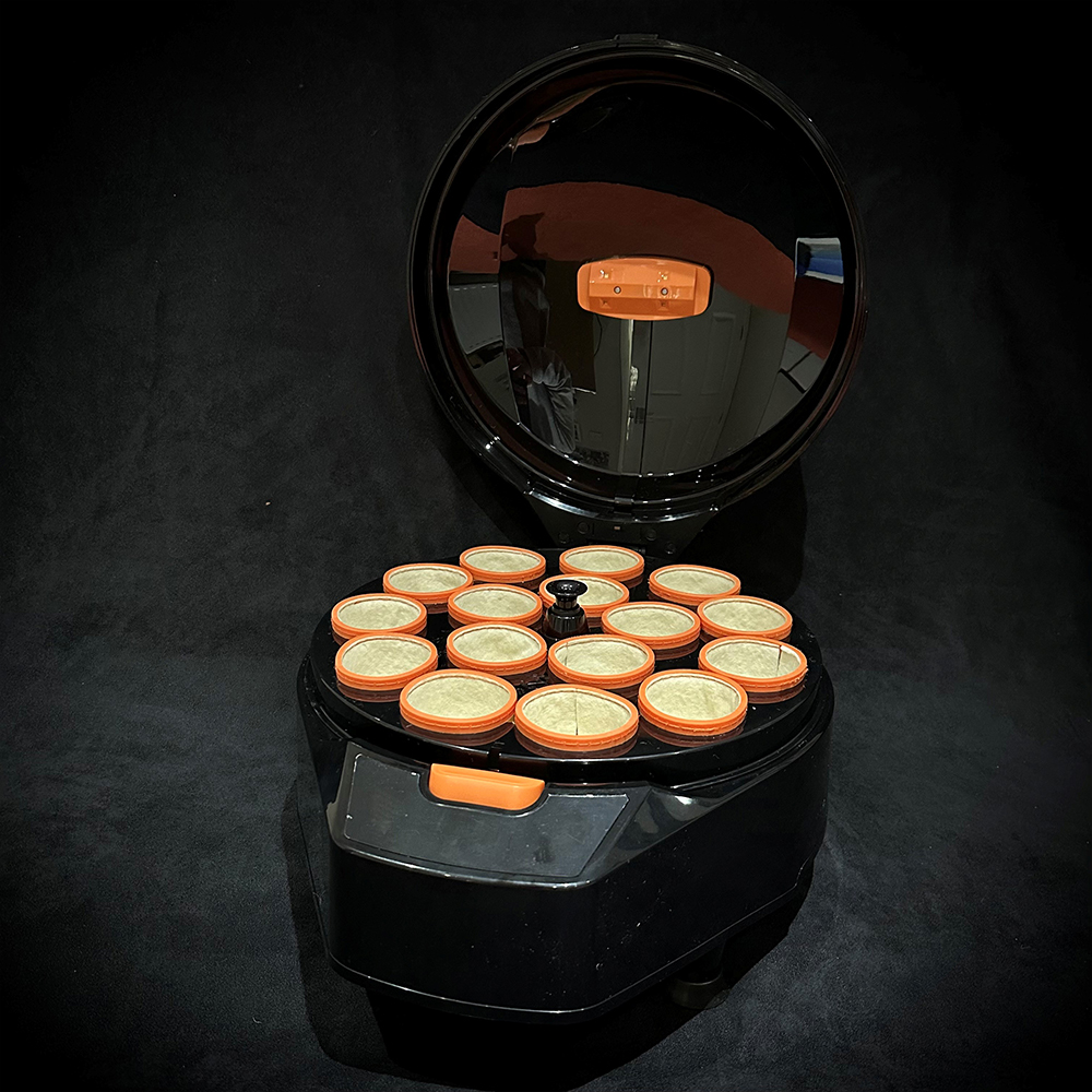 Schwarze Billard-Kugelpoliermaschine GEN 5 mit offenem, glänzendem Deckel. Innen sind 16 runde Reinigungsbecher aus hellem Wollmaterial mit orangefarbenen Rändern kreisförmig angeordnet. Drehknopf mittig, orange Taste vorn.