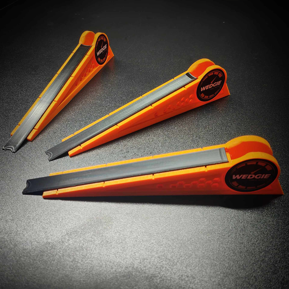 Three identical, bright orange and black, wedge-shaped pool table testers lie diagonally on a dark, slightly textured surface. Each wedge has a black indentation and a round ‘WEDGIE’ logo with a speedometer.