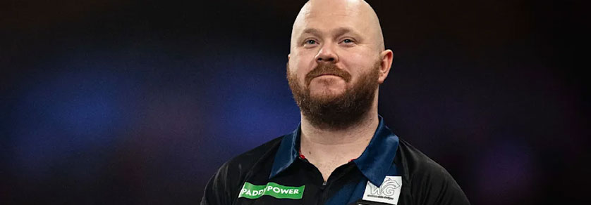 Un homme chauve, à la barbe brun-rougeâtre et au sourire discret, regarde vers la droite. Il porte un polo noir rehaussé d’un col bleu et arborant le logo vert « Paddy Power ». L’arrière-plan, sombre et flou, présente de légers reflets bleus.