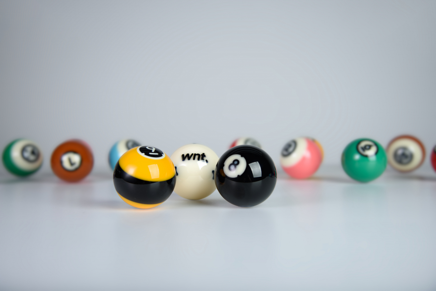 Glänzende Poolbillard-Kugeln der Dynaspheres Titan Serie auf hellem Untergrund. Im Fokus: gelb-schwarz gestreifte Neun, weiße Spielkugel mit WNT-Logo und schwarze Acht. Im Hintergrund weitere bunte Kugeln.