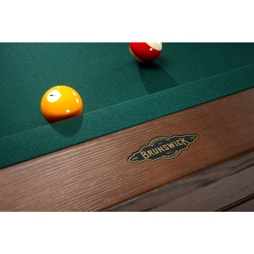 Close-up of the Brunswick Winfield pool table with dark green cloth. An orange ball lies at the front, a red and white striped one at the back. The wooden rails, finished in nutmeg, feature a fine grain and the golden ‘ Brunswick ’ emblem.