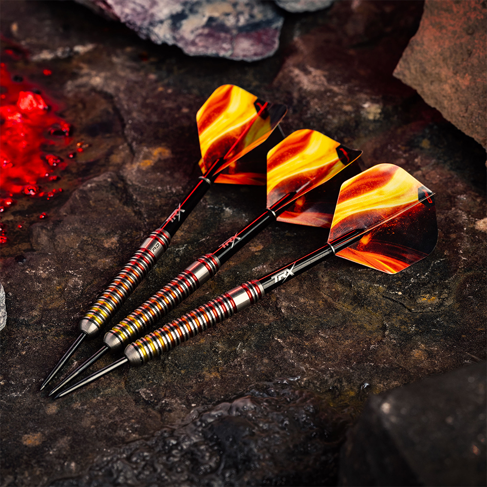 Three steel darts with flame-like red and yellow flights lie diagonally on dark, rough rock. Finely ribbed metal barrels with gold and red accents in the ‘Furnace’ design. Black tips point downwards. Red splashes on the left.